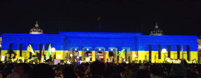 das Heldentor am Wiener Heldenplatz in den Farben der Urkraine bestrahlt. Davor Menschen und Fahnen.